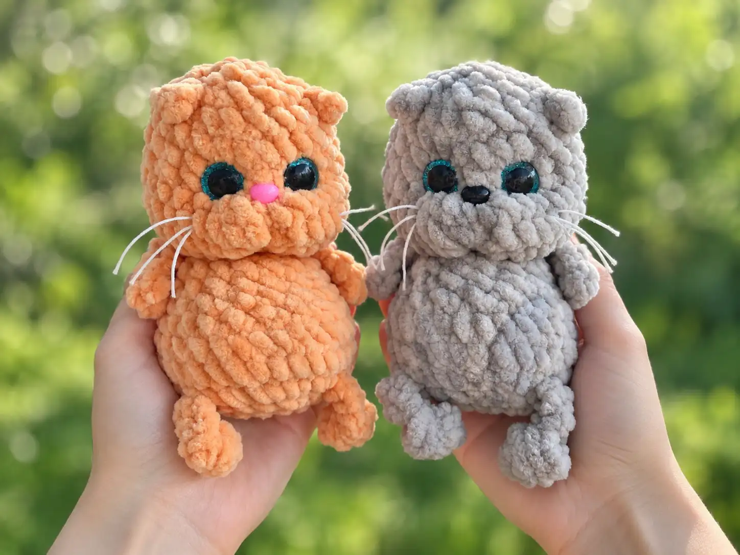 Two hand-held crochet kittens, one orange and one gray, outdoors near a potted plant, with visible whisker details.
