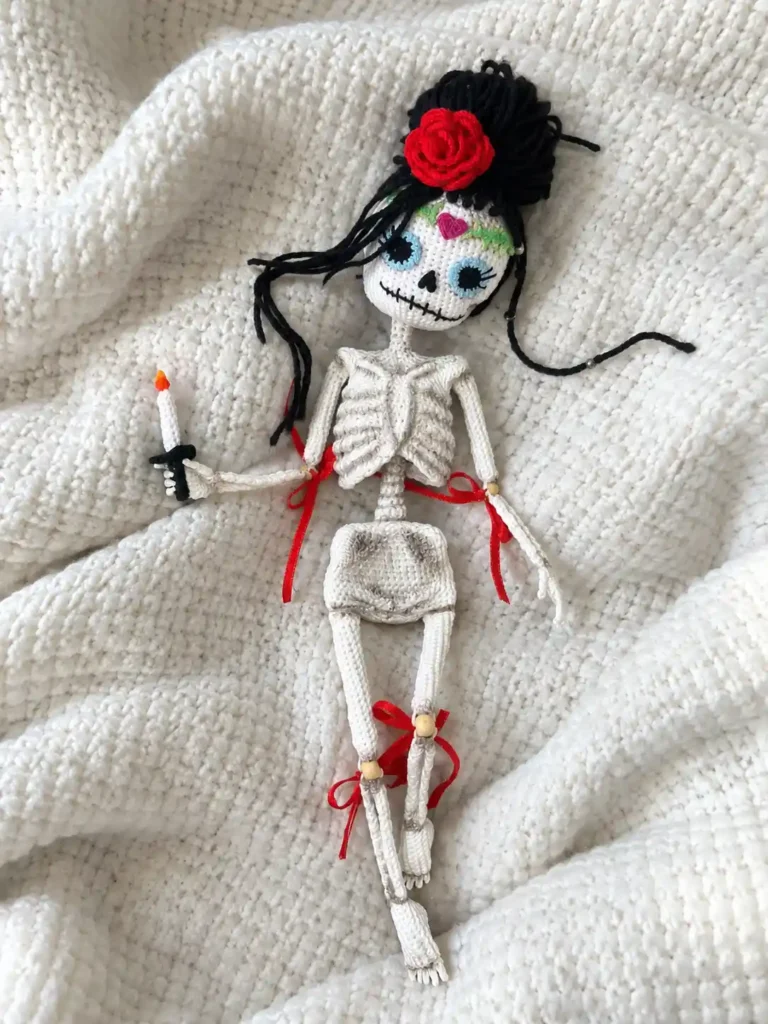 White yarn skeleton doll crochet pattern with red joint ribbons and a hair rose, lying flat on a textured white blanket.