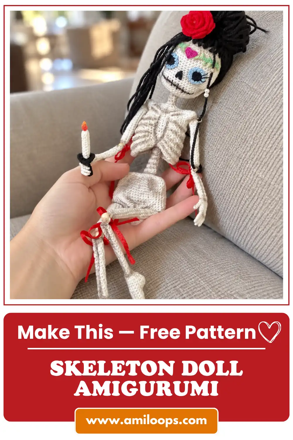 Hand holding a white yarn skeleton doll crochet pattern with red ribbons and a red hair rose against a soft grey couch.