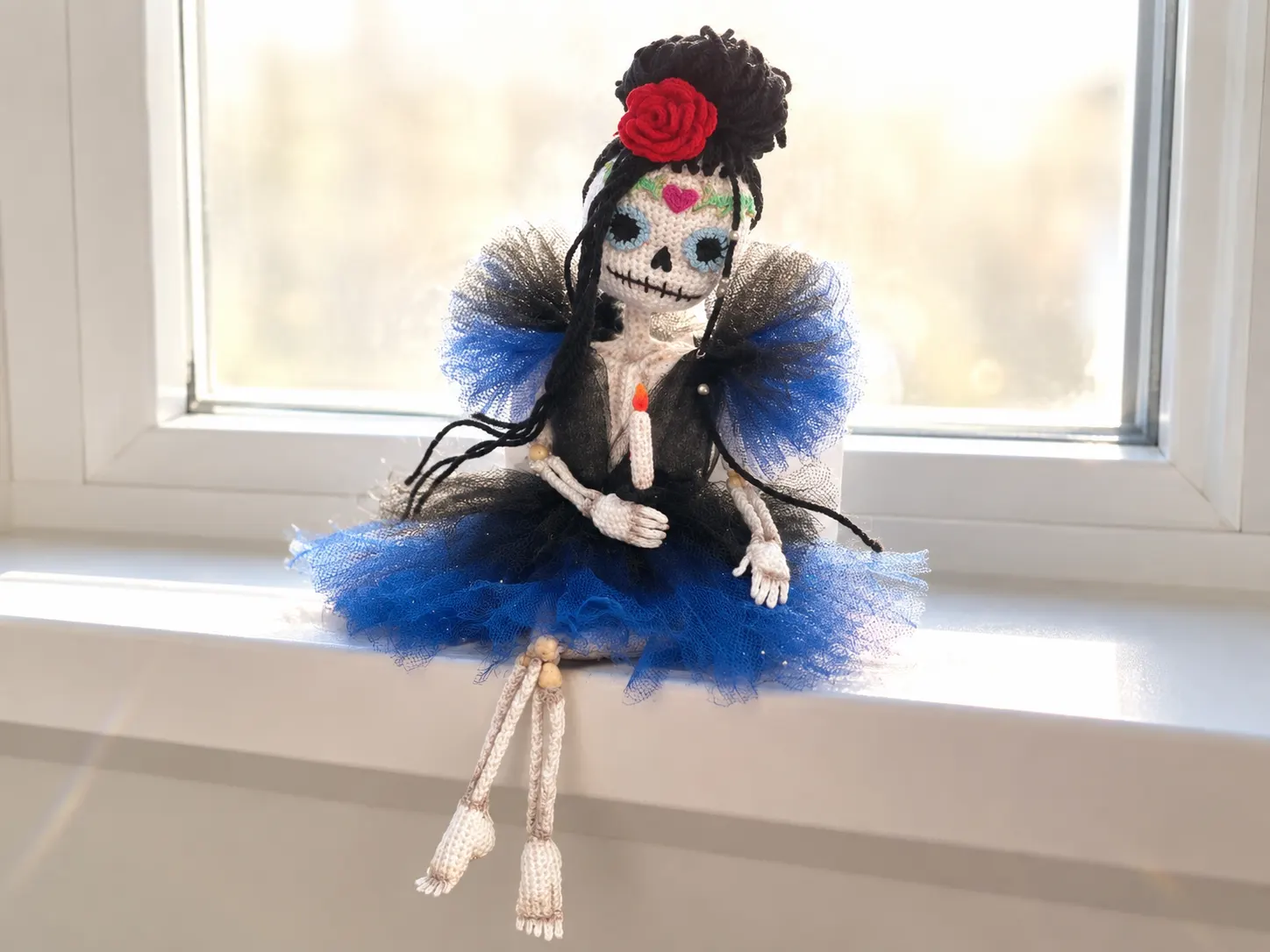White yarn skeleton doll crochet pattern in a blue tulle dress with a red rose, sitting on a sunny white windowsill.