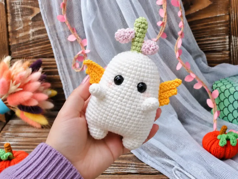 Hand holding a white bulb amigurumi pattern toy with yellow wings against rustic wood and blue fabric.