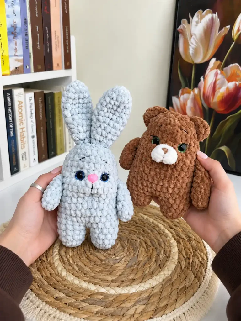 Hands holding light blue bunny and brown bear plush toys made with soft chenille yarn, displayed in front of a white bookshelf and tulip painting.