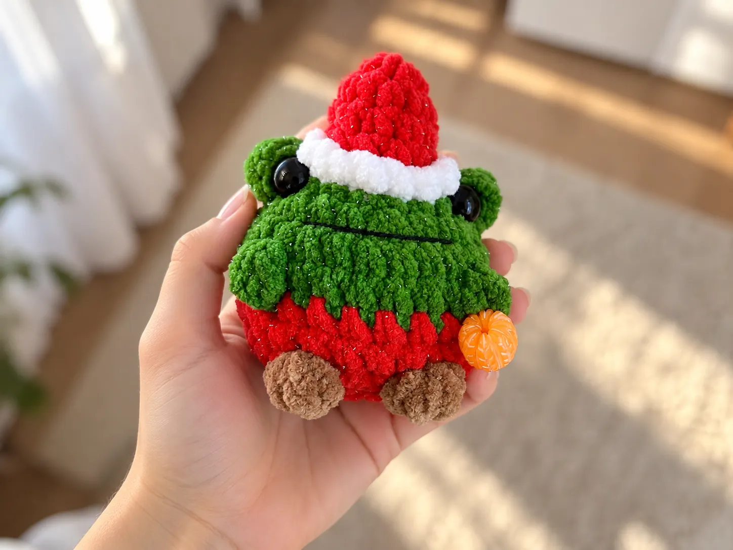 Sparkly green plush crochet frog in a hat, wearing a red santa suit, held in a hand indoors