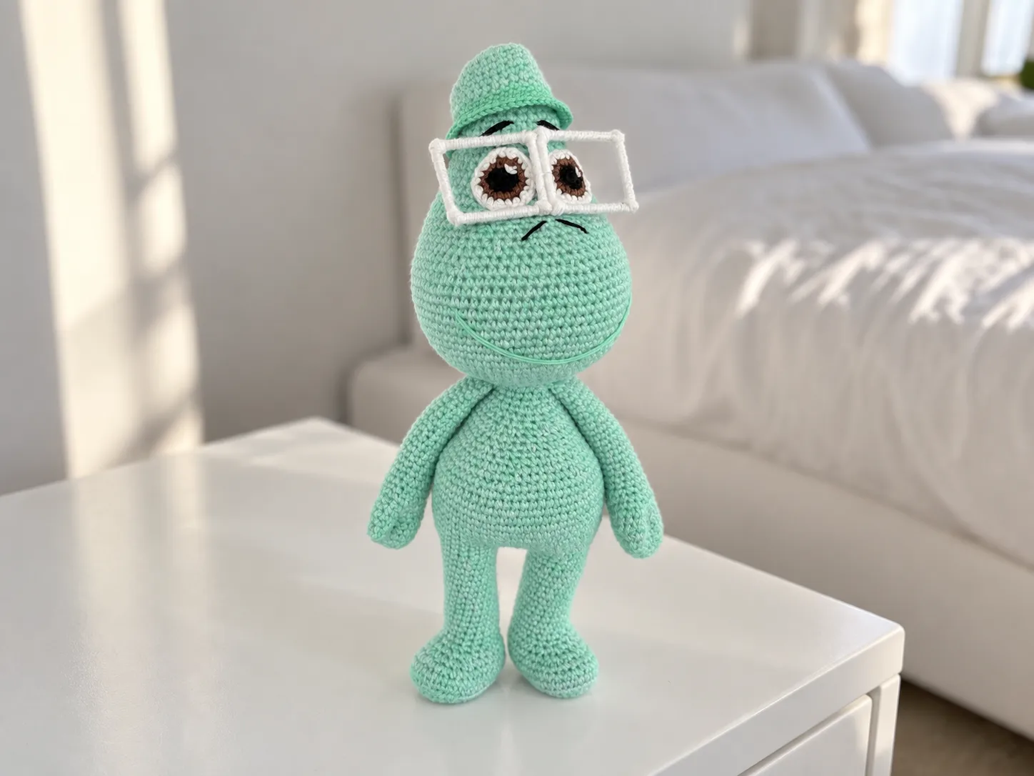 Mint green joe gardner amigurumi toy with white glasses and hat standing on a white nightstand in a bright bedroom