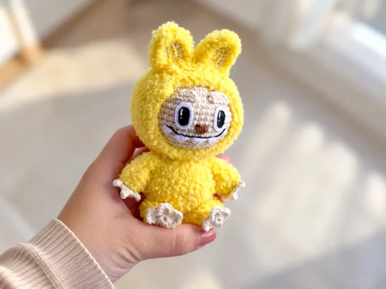 A hand holding a fluffy yellow and beige amigurumi toy with cute teeth, displayed indoors with natural daylight.