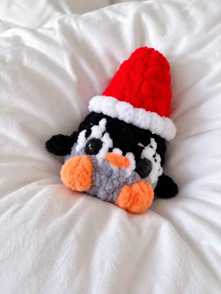 Black, white, and grey plush yarn penguin with an orange beak and feet, wearing a red and white Santa hat on white bedding.