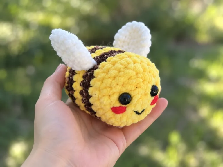 Hand holding a yellow and brown plush bee amigurumi toy with white wings, black safety eyes, and red cheeks against a green background.